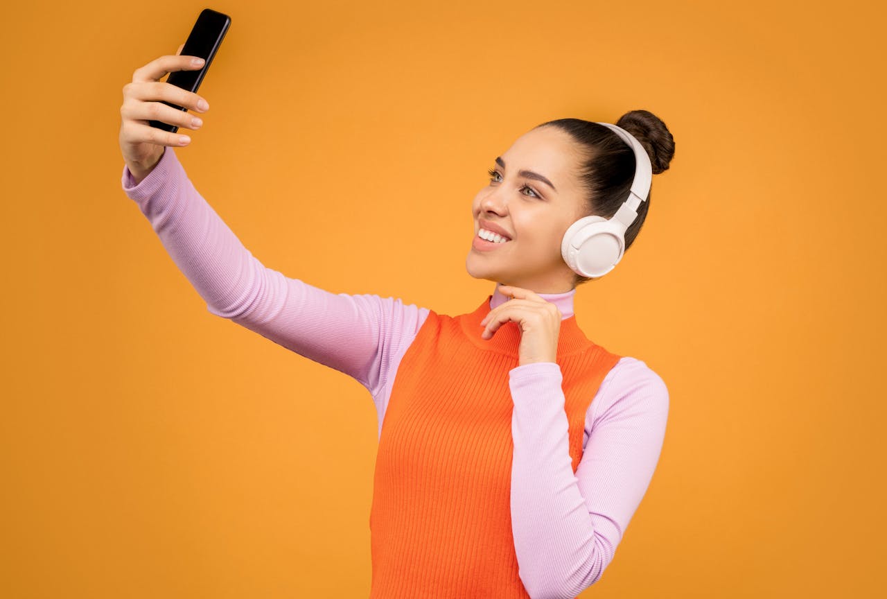 Portrait of a young woman with headphones taking a selfie against an orange background.
